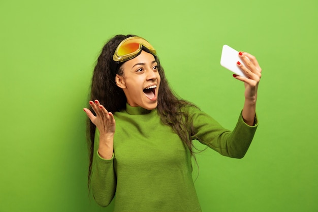 Retrato de mujer morena joven afroamericana en pasamontañas sobre fondo verde de estudio. Concepto de emociones humanas, expresión facial, ventas, publicidad, deportes de invierno y vacaciones. Haciendo selfies o vlogs.