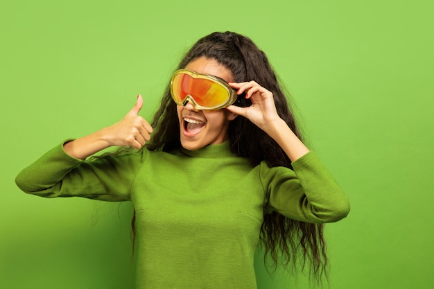 Retrato de mujer morena joven afroamericana en pasamontañas sobre fondo verde de estudio. Concepto de emociones humanas, expresión facial, ventas, publicidad, deportes de invierno y vacaciones. Sonriendo, pulgar hacia arriba.