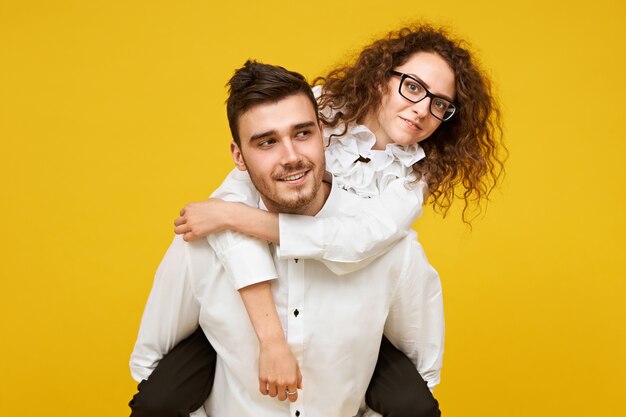 Retrato de mujer morena joven feliz en anteojos disfrutando de un paseo a cuestas en la espalda de su novio contra la pared amarilla en blanco. Concepto de amor, romance, unión y felicidad.