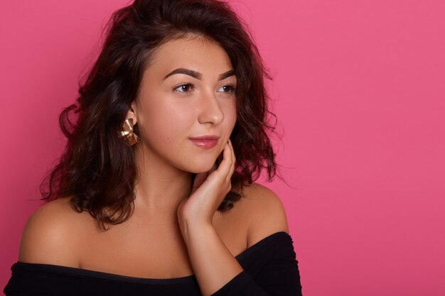 Retrato de mujer morena joven maravillosa mirando soñadoramente a un lado, manteniendo la mano en el cuello, posando con los hombros desnudos, chica de cabello oscuro con aretes.