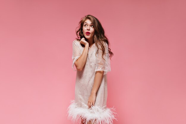 Retrato de mujer morena rizada en vestido blanco posando de buen humor en la pared rosa