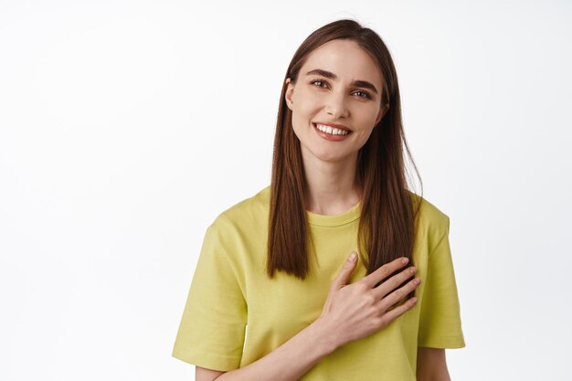 Retrato de mujer morena sonriente decir gracias, apreciar algo, tomar la mano en el corazón, apreciar el cumplido, ser halagada, de pie contra el fondo blanco