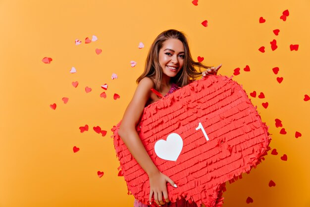 Retrato de mujer morena sonriente obsesionada con las redes sociales. Retrato de interior de niña glamorosa de pie sobre amarillo bajo confeti.