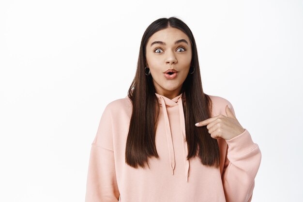 Retrato de mujer morena sorprendida y emocionada jadeando, apuntando a sí misma con asombro, ganando el premio, siendo elegida, de pie contra la pared blanca