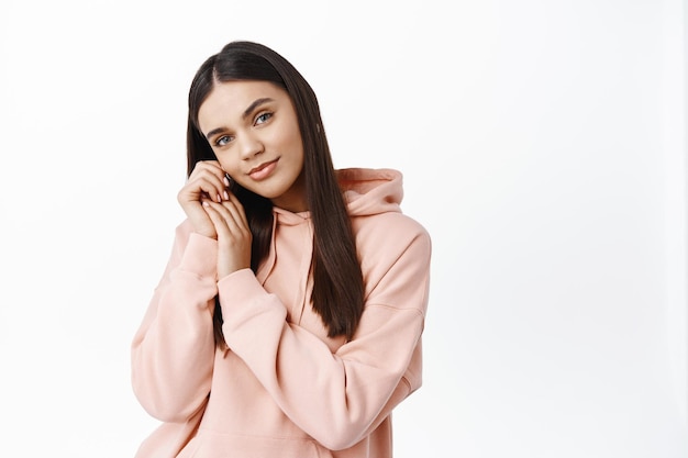 Retrato de mujer morena tierna y romántica, apoyándose suavemente en las manos y sonriendo sensualmente al frente, de pie en una sudadera con capucha contra la pared blanca