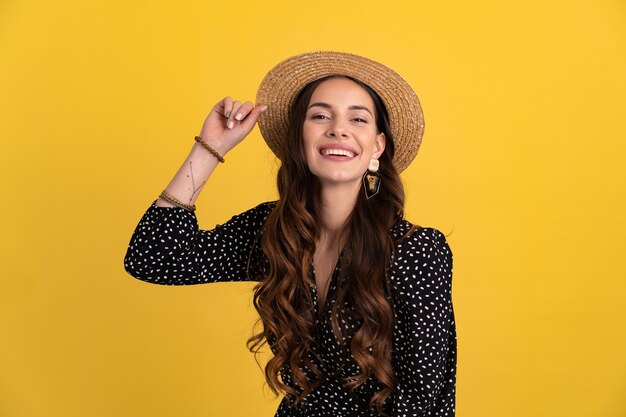 Retrato de mujer muy atractiva posando aislada sobre fondo amarillo con vestido negro punteado y sombrero de paja