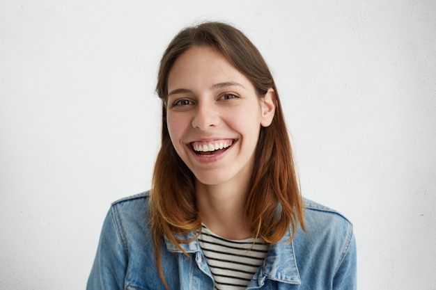 Retrato de mujer natural positiva con cabello oscuro en chaqueta vaquera sonriendo ampliamente