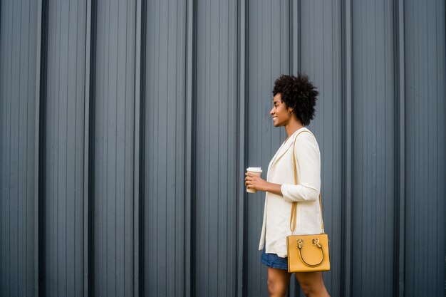 Retrato de mujer de negocios afro sosteniendo una taza de café mientras camina al aire libre en la calle. Concepto urbano y empresarial.