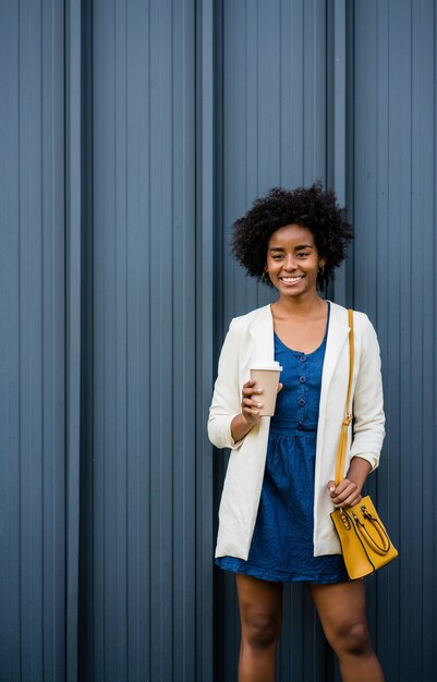 Retrato de mujer de negocios afro sosteniendo una taza de café mientras está parado al aire libre en la calle. Concepto urbano y empresarial.