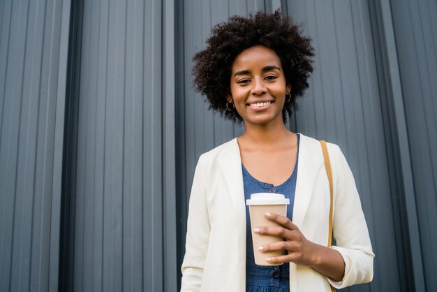 Retrato de mujer de negocios afro sosteniendo una taza de café mientras está de pie al aire libre en la calle