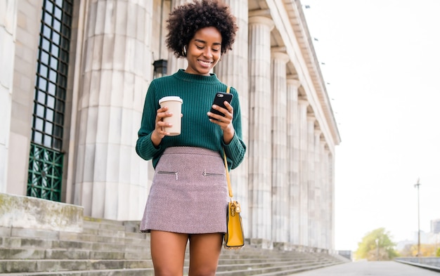 Retrato de mujer de negocios afro usando su teléfono móvil y sosteniendo una taza de café mientras camina al aire libre en la calle