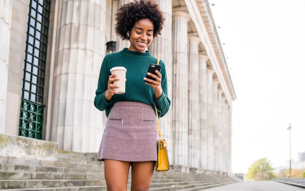 Retrato de mujer de negocios afro usando su teléfono móvil y sosteniendo una taza de café mientras camina al aire libre en la calle