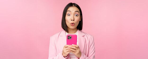 Retrato de mujer de negocios asiática con cara sorprendida usando una aplicación de teléfono inteligente con traje de negocios Chica coreana con teléfono móvil y expresión de cara emocionada fondo rosa