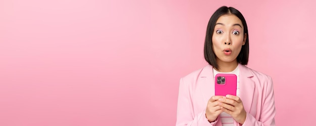 Retrato de mujer de negocios asiática con cara sorprendida usando una aplicación de teléfono inteligente con traje de negocios Chica coreana con teléfono móvil y expresión de cara emocionada fondo rosa