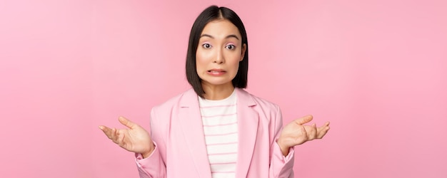 Retrato de una mujer de negocios asiática confundida encogiéndose de hombros que parece despistada y desconcertada, no sé, no puedo decir que esté de pie sobre un fondo rosa en traje de oficina