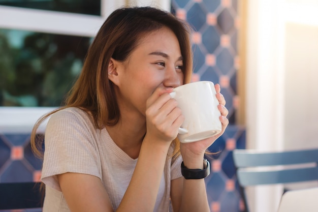Retrato de la mujer de negocios asiática joven feliz con la taza en manos que bebe el café por la mañana