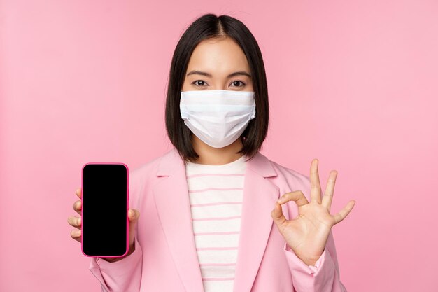 Retrato de una mujer de negocios asiática con mascarilla médica covid19 que muestra la pantalla del teléfono inteligente y el fondo rosa de la señal OK