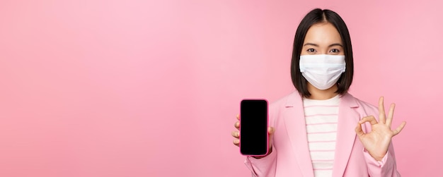 Retrato de una mujer de negocios asiática con mascarilla médica covid19 que muestra la pantalla del teléfono inteligente y el fondo rosa de la señal OK