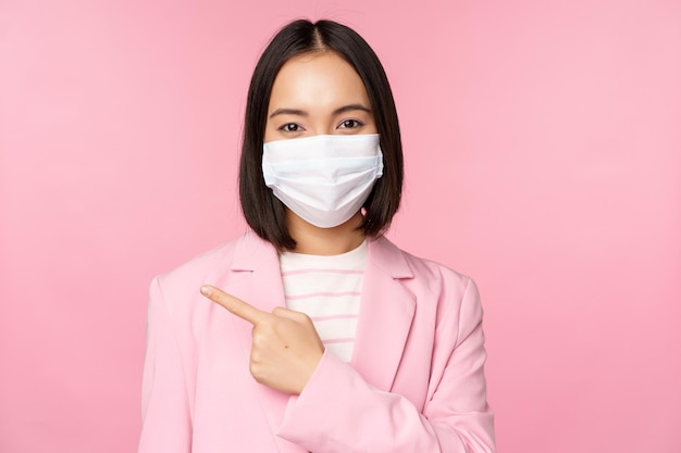 Retrato de una mujer de negocios asiática con mascarilla médica y traje que señala con el dedo a la izquierda mostrando el fondo rosa del estudio de banner de la empresa de publicidad