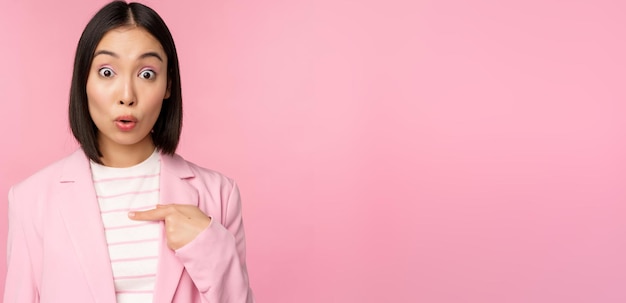 Retrato de mujer de negocios asiática reacciona sorprendida de sí misma con incredulidad en la cara posando en traje contra fondo rosa