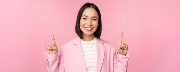 Retrato de una mujer de negocios asiática señalando con el dedo hacia arriba y sonriendo mostrando la información del logotipo de la empresa en la parte superior de pie sobre un fondo rosa