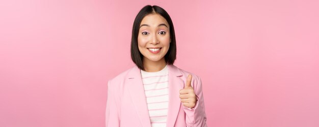 Retrato de una mujer de negocios asiática sonriendo satisfecha mostrando los pulgares hacia arriba alabando como y aprobando estar de pie en traje sobre fondo rosa