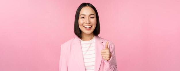 Retrato de una mujer de negocios asiática sonriendo satisfecha mostrando los pulgares hacia arriba alabando como y aprobando estar de pie en traje sobre fondo rosa