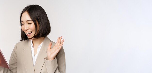 Retrato de una mujer de negocios asiática sonriente chateando en video en una tableta digital agitando la mano en la pantalla del dispositivo de pie en traje sobre fondo blanco