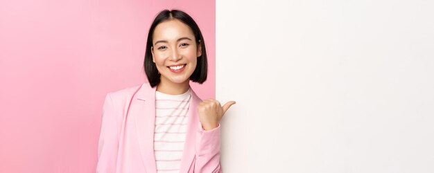 Retrato de mujer de negocios asiática sonriente en traje dama corporativa señalando con el dedo a la pared vacía blanca b