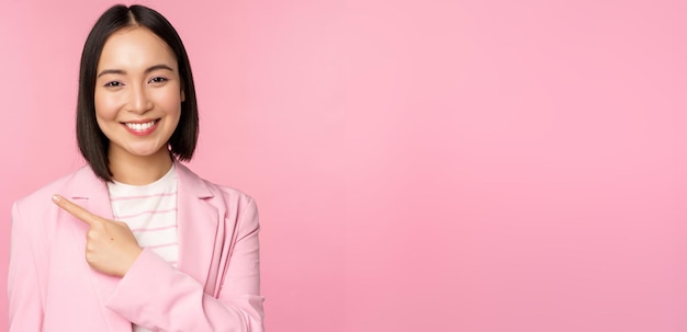 Retrato de mujer de negocios asiática vendedora en traje señalando con el dedo a la izquierda mostrando un anuncio de pancarta sonriendo y con un fondo rosa profesional