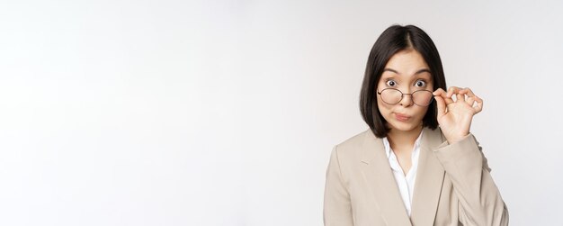 Retrato de mujer de negocios despegando gafas y mirando sorprendida a la cámara de pie sobre fondo blanco.