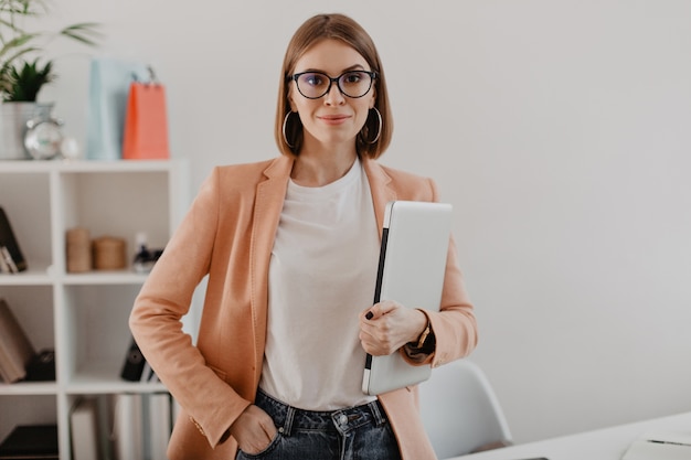 Retrato de mujer de negocios exitosa con gafas y chaqueta ligera sonriendo contra la oficina blanca.