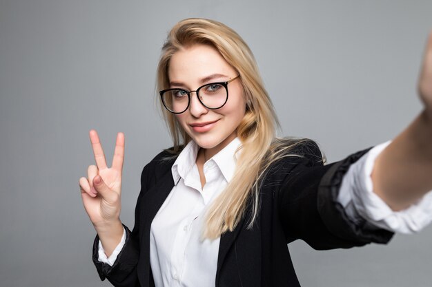 Retrato de mujer de negocios haciendo selfie usando teléfono inteligente e internet, gesticulando símbolo de paz, con videollamada en línea sobre pared gris