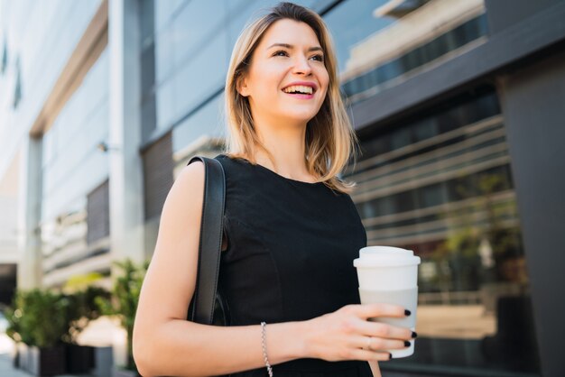 Retrato de mujer de negocios joven caminando al trabajo mientras bebe café para llevar. Concepto de negocio y éxito.