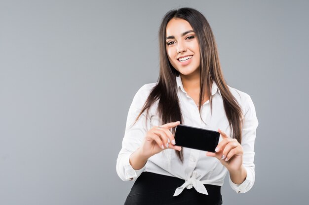 Retrato de una mujer de negocios joven emocionada que señala el teléfono móvil de la pantalla en blanco aislado en blanco