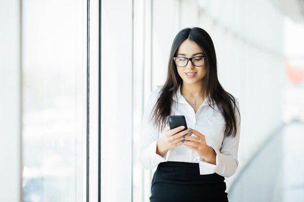 Retrato de una mujer de negocios joven escribiendo teléfono de texto contra ventanas panorámicas. Concepto de negocio