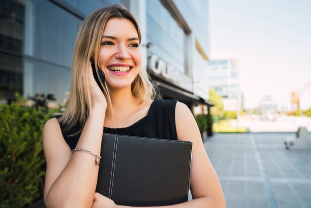 Retrato de mujer de negocios joven hablando por teléfono mientras está de pie fuera de los edificios de oficinas. Concepto de negocio y éxito.