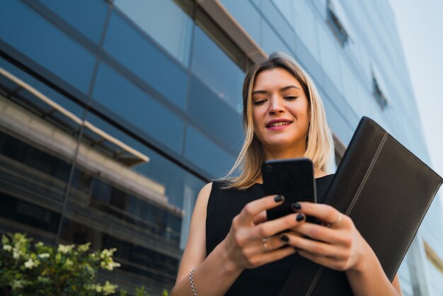 Retrato de mujer de negocios joven que usa su teléfono móvil mientras está de pie fuera de los edificios de oficinas. Concepto de negocio y éxito.