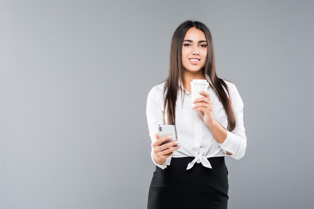 Retrato de una mujer de negocios joven satisfecha con teléfono móvil mientras sostiene una taza de café para ir aislado en blanco