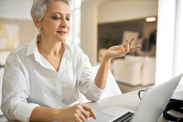 Retrato de mujer de negocios madura exitosa enérgica con camisa blanca que tiene una reunión de negocios en línea a través de una videoconferencia, gesticulando emocionalmente, discutiendo el acuerdo