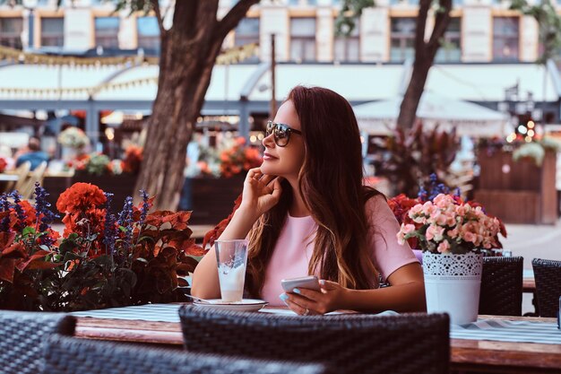 Retrato de una mujer de negocios de mediana edad con cabello castaño largo con gafas de sol sostiene un teléfono inteligente mientras se sienta en el café al aire libre.