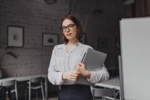 Retrato de mujer de negocios de ojos marrones en traje blanco y negro y elegantes gafas posando con laptop en sala blanca.