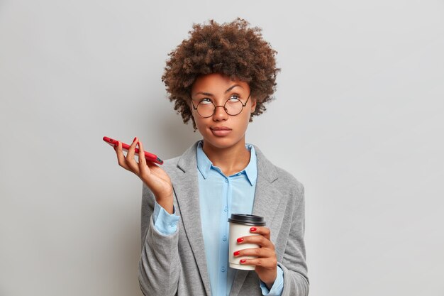 Retrato de mujer de negocios pensativa con cabello afro tiene expresión aburrida espera llamada tiene teléfono móvil