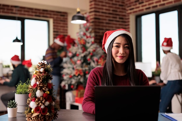 Retrato de una mujer de negocios que trabaja en la temporada navideña en una oficina de inicio decorada con adornos y luces navideñas festivas. Trabajador de una empresa asiática que usa una computadora portátil durante la celebración de la temporada.