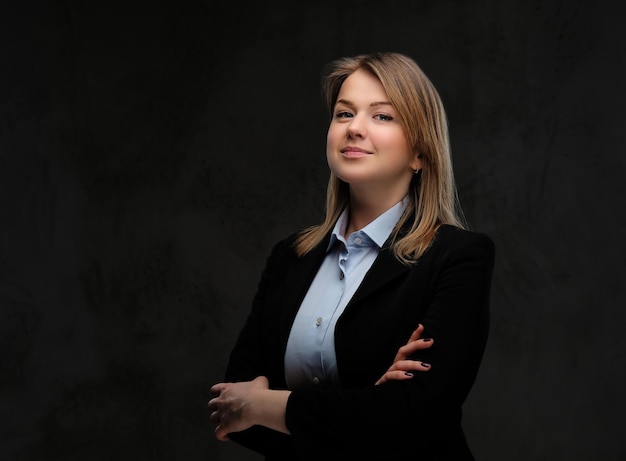 Retrato de una mujer de negocios rubia sonriente vestido formal de pie con los brazos cruzados. Aislado en un fondo de textura oscura.