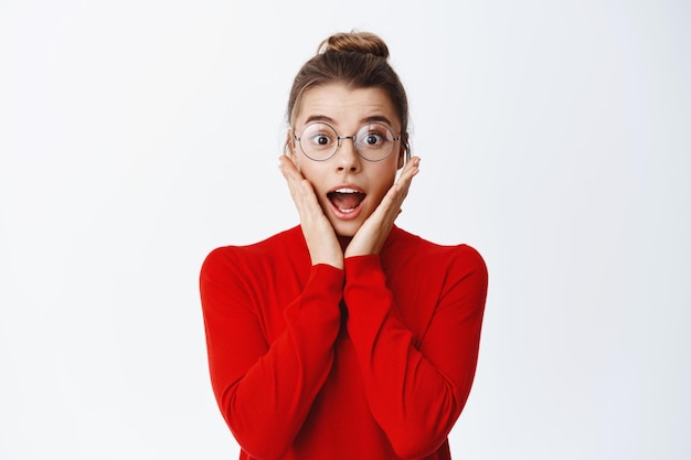 Retrato de mujer de negocios rubia sorprendida con gafas, jadeando y con la mandíbula caída asombrada, mirando algo genial, tomándose las manos en las mejillas y mirando impresionado en blanco