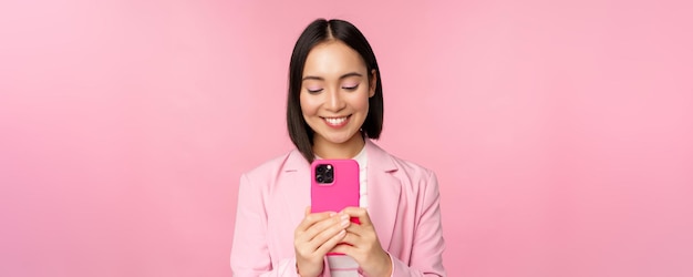 Retrato de una mujer de negocios sonriente persona corporativa asiática que usa una aplicación de teléfono móvil para teléfonos inteligentes de pie sobre un fondo rosa