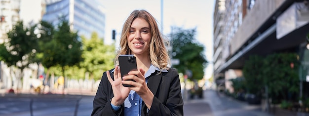 Retrato de una mujer de negocios sonriente usando un teléfono móvil mientras está de pie al aire libre cerca de edificios de oficinas