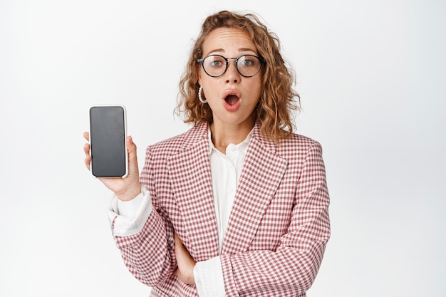 Retrato de mujer de negocios sorprendida que muestra la pantalla del teléfono inteligente, jadeando de asombro, mirando impresionado en blanco