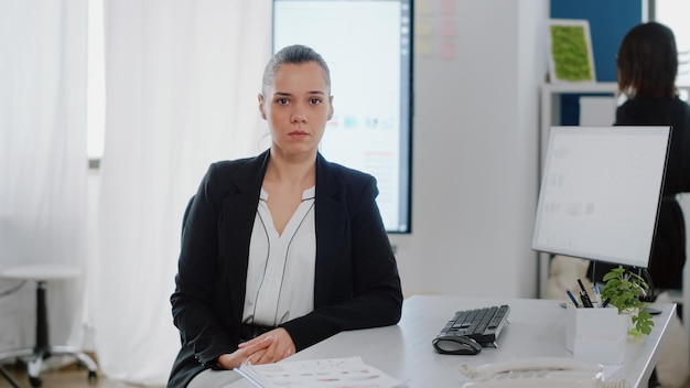 Retrato de mujer de negocios con trabajo de oficina sentada en el escritorio con monitor de computadora y gráficos de datos en pantalla. Empleado corporativo mirando la cámara y preparándose para trabajar en un proyecto empresarial